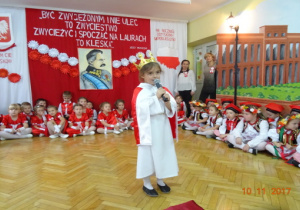 Na tle biało czerwonej dekoracji stoi dziewczynka przebrana za Polskę.